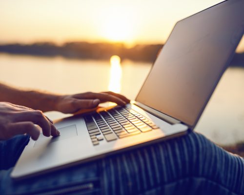 Close up of Caucasian man sitting in nature and using laptop. In background is river and sunset. Selective focus on hands.